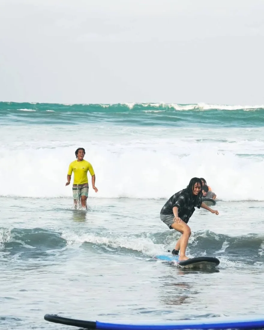 A student standing up on her board at Sabang Beach while her El Dawn instructor cheers behind her